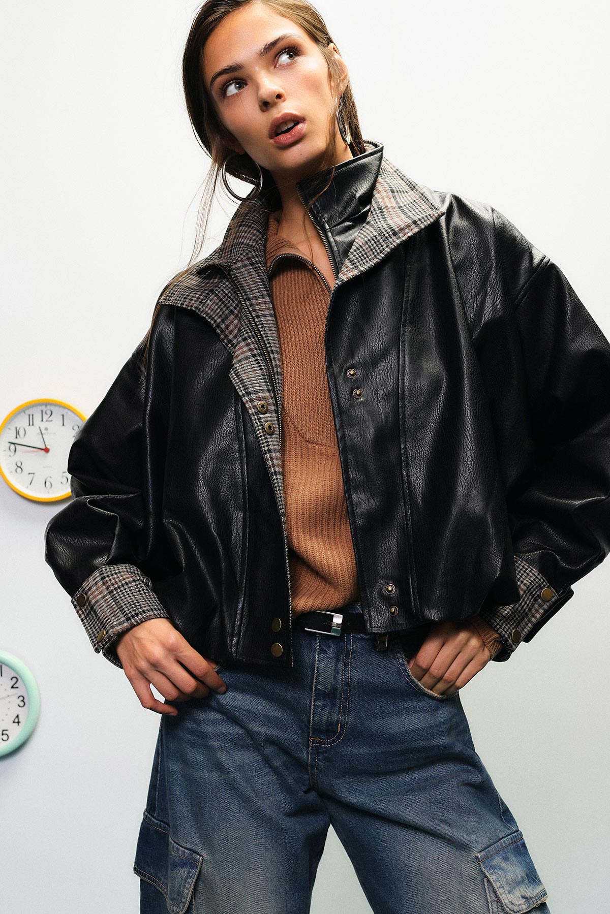 Young woman wearing a black leather jacket, brown sweater and blue jeans, posing with hands on hips against a white wall with clocks