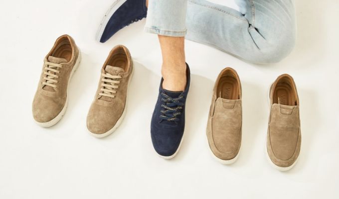 Person wearing a navy casual sneaker among four tan suede casual shoes on a white background, jeans visible