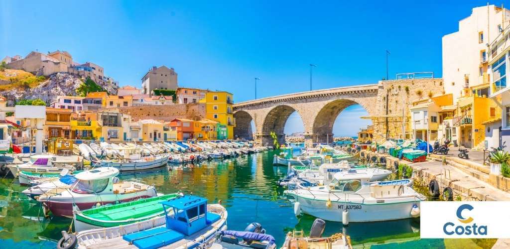 Colourful Mediterranean harbour with moored fishing boats, stone arched bridge and pastel coastal houses under clear blue sky