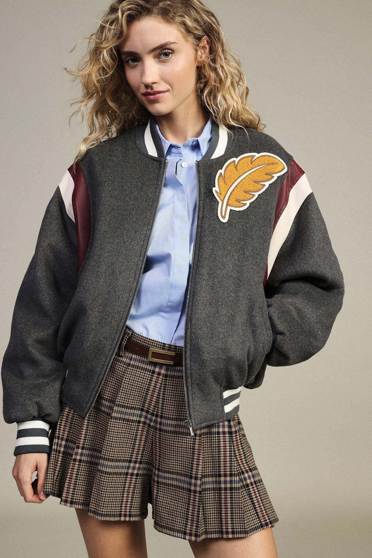 Young woman wearing a grey varsity jacket with a yellow leaf patch, blue shirt and pleated plaid skirt against a neutral background