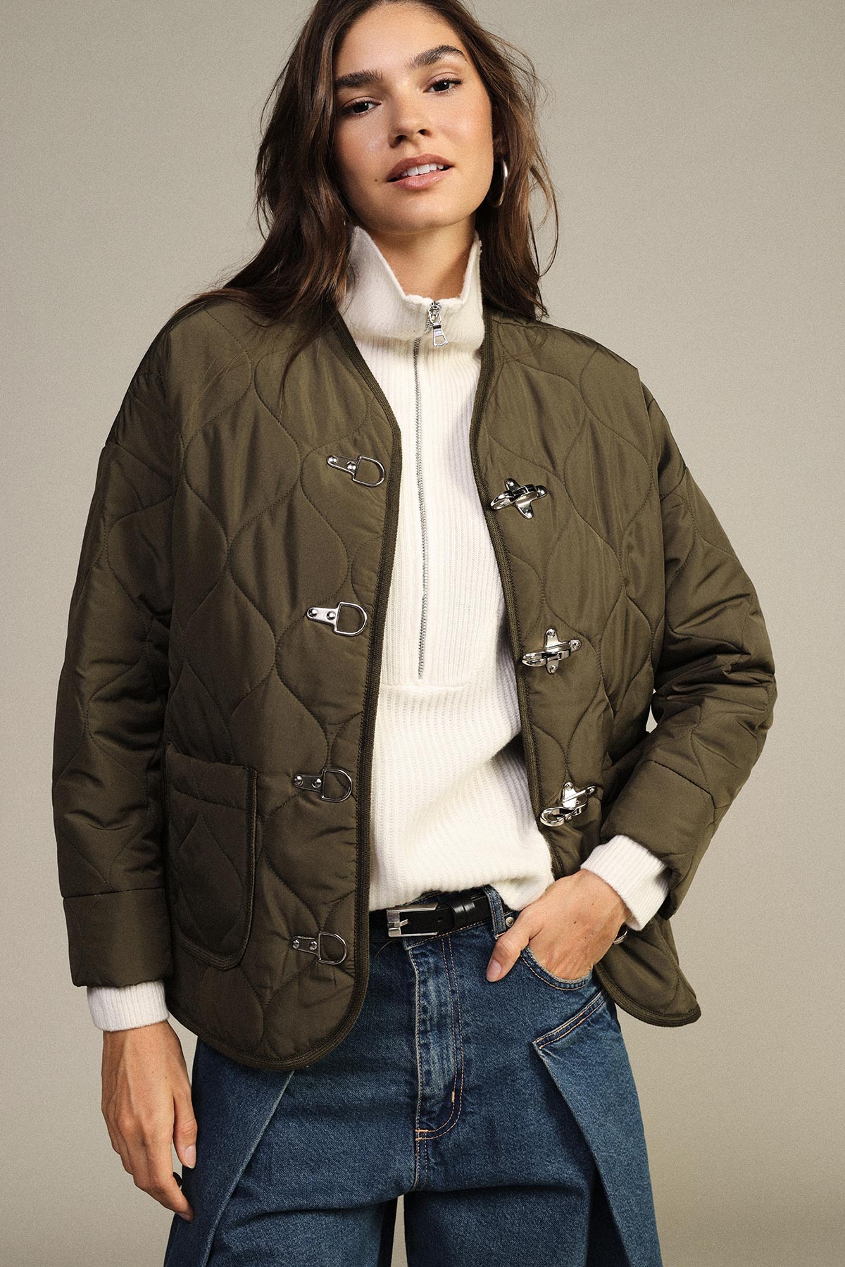 Person wearing a quilted olive-green jacket with metal toggle closures over a cream zip-up sweater and blue jeans