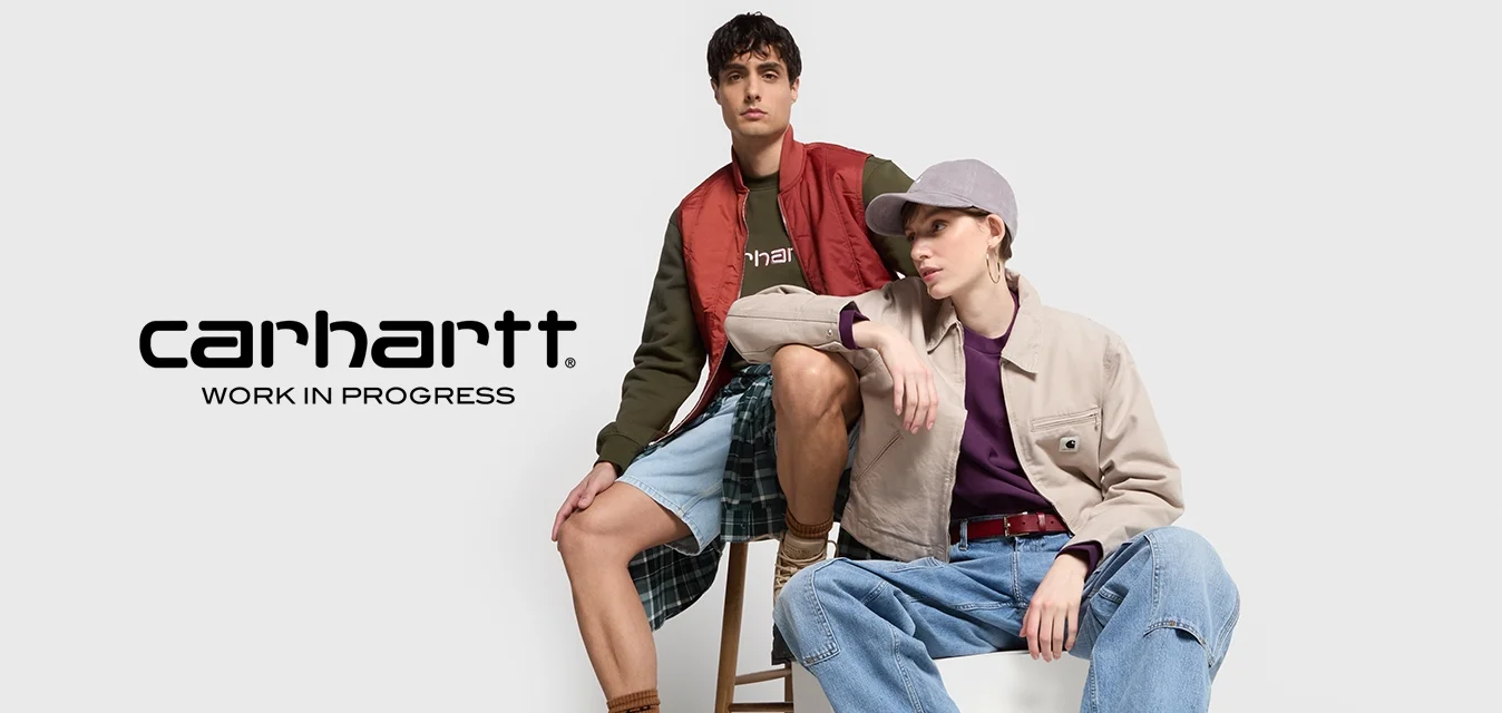 Due modelli indossano outfit casual a strati. Uno con un gilet imbottito rosso e una felpa verde scuro, l'altro con una giacca beige sopra un top bordeaux.