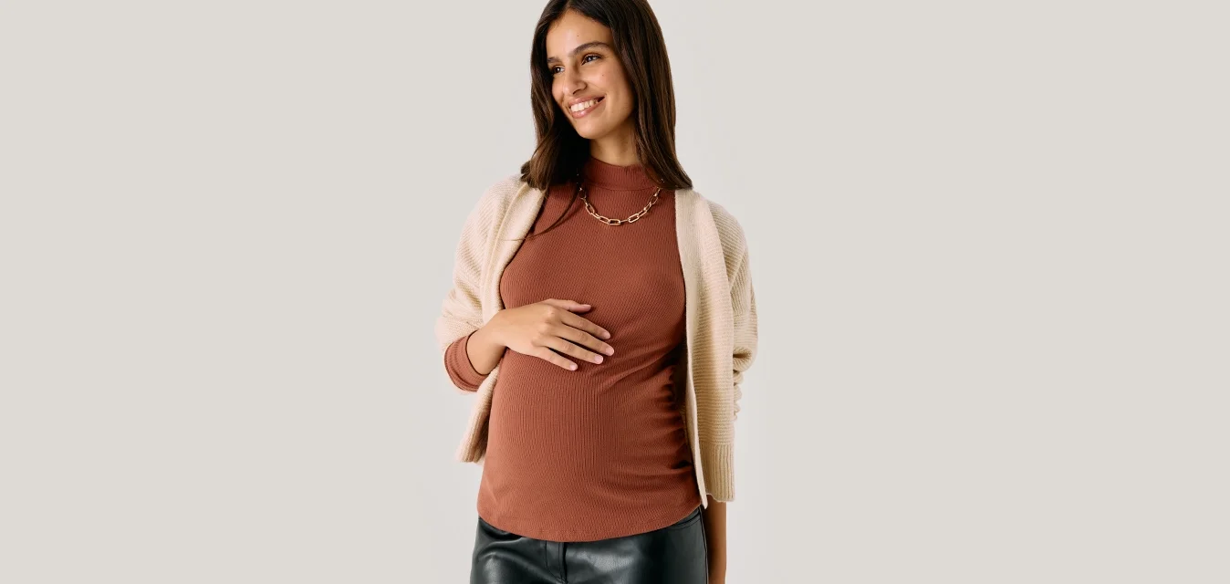 Donna incinta sorridente che tiene la pancia, indossando un top di maglia marrone, un cardigan beige, pantaloni neri e una collana d'oro, in piedi su uno sfondo semplice.
