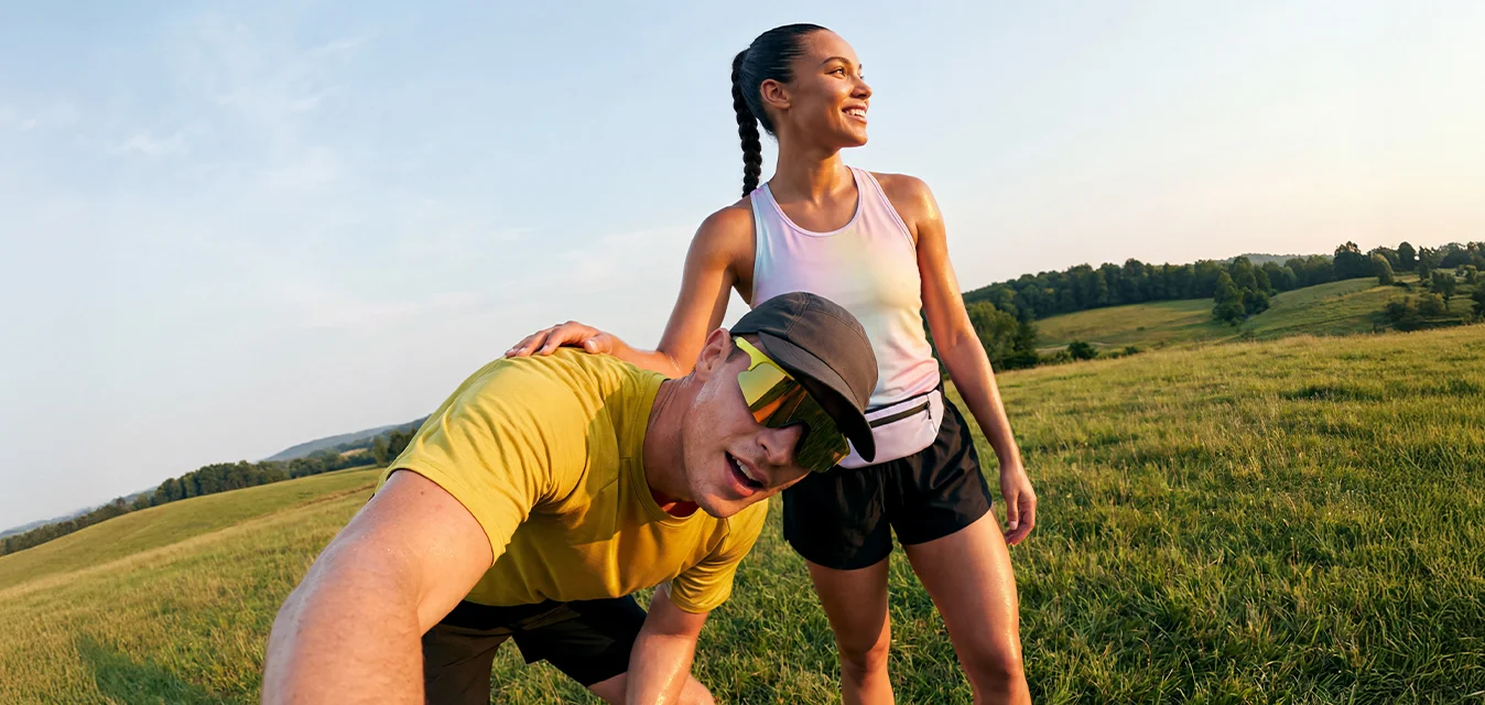 Due corridori si riposano su un campo erboso al tramonto, uno fa un selfie mentre l'altro sorride, entrambi vestiti con abbigliamento sportivo.