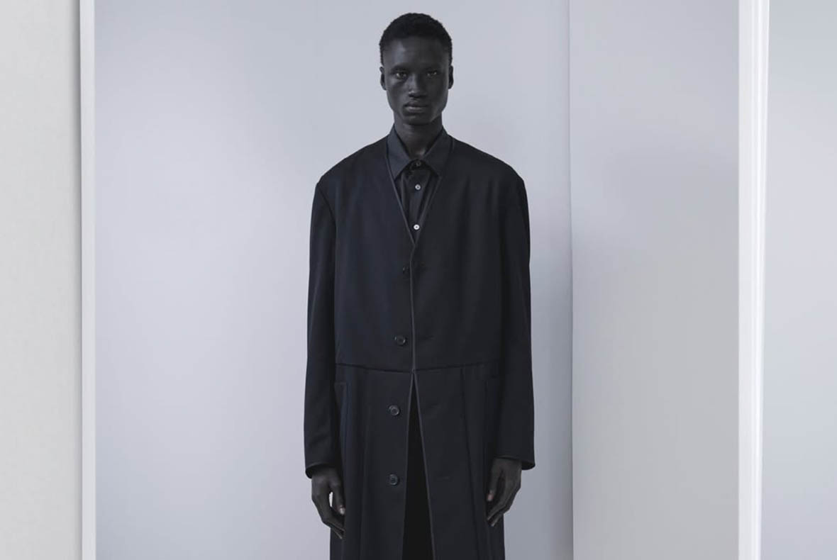 Model in minimalist studio wearing long black buttoned coat and shirt, contemporary men's fashion portrait