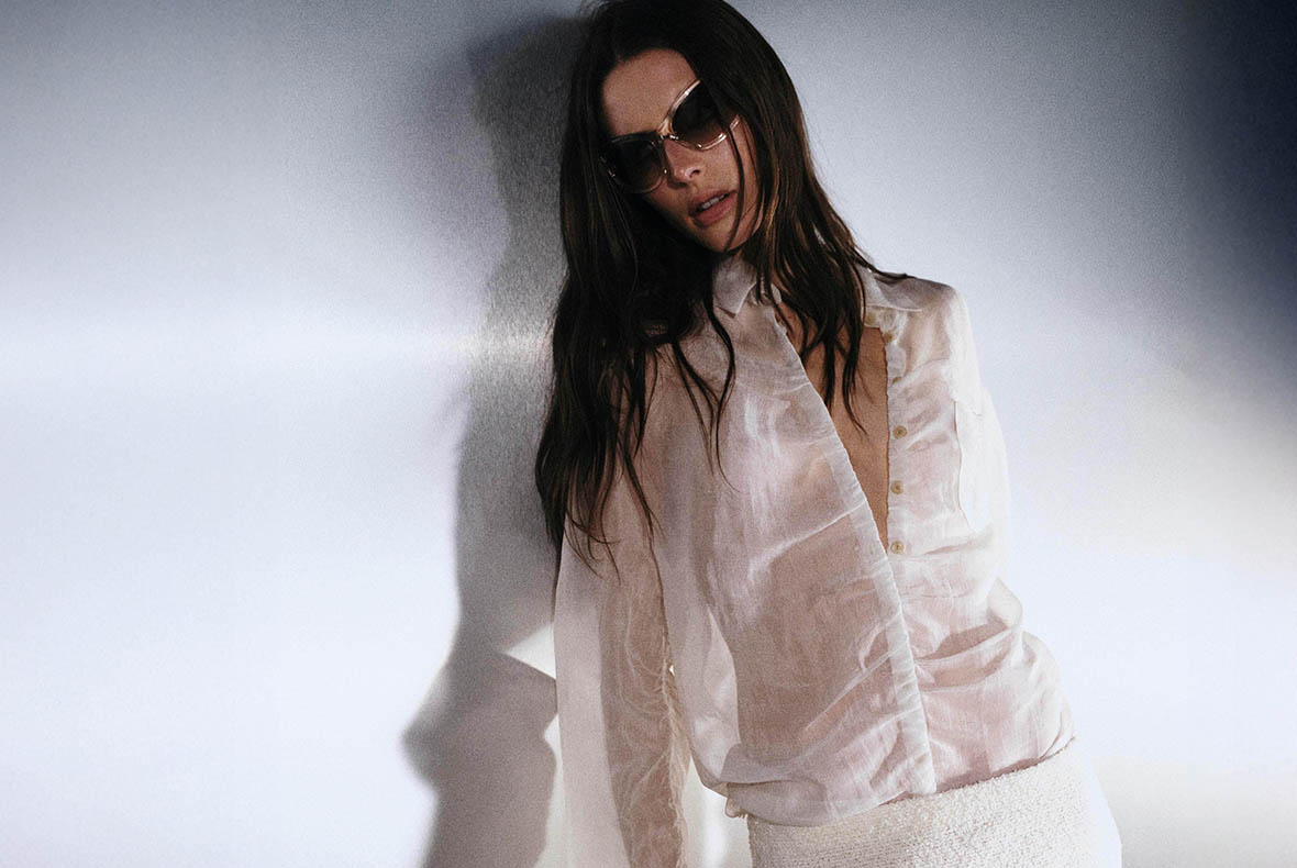 Fashion portrait of a woman in sunglasses wearing a sheer white blouse and textured skirt, leaning against a light wall with soft shadows