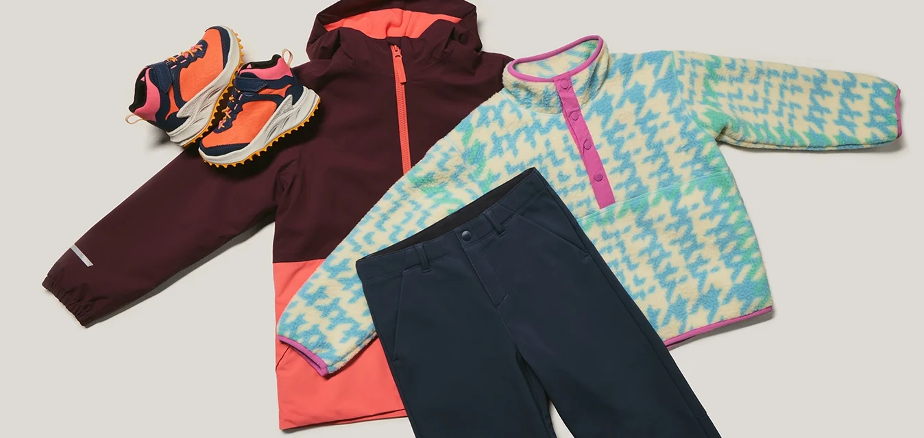 Outfit invernale per bambini composto da scarpe da trekking arancioni e viola, giacca bordeaux e corallo, felpa in pile blu a fantasie e pantaloni blu navy disposti in piano.