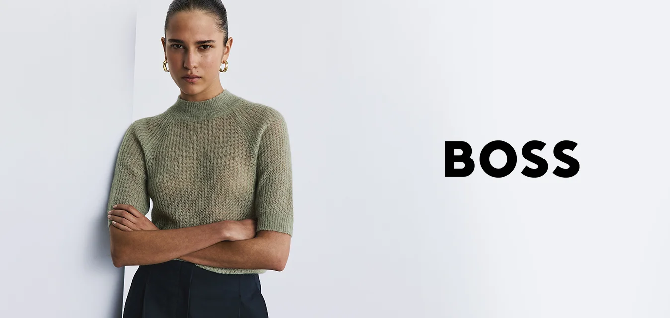 Giovane donna con i capelli raccolti dietro, indossa orecchini a cerchio dorati, un maglione lavorato a maglia verde a maniche corte e pantaloni scuri, è in piedi con le braccia incrociate.