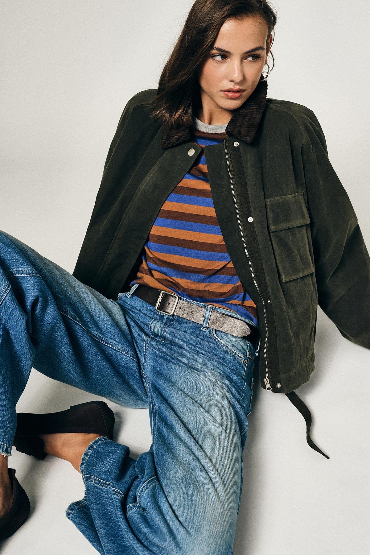 Woman in olive suede jacket, brown-and-blue striped top and relaxed blue jeans seated in casual women's fashion pose