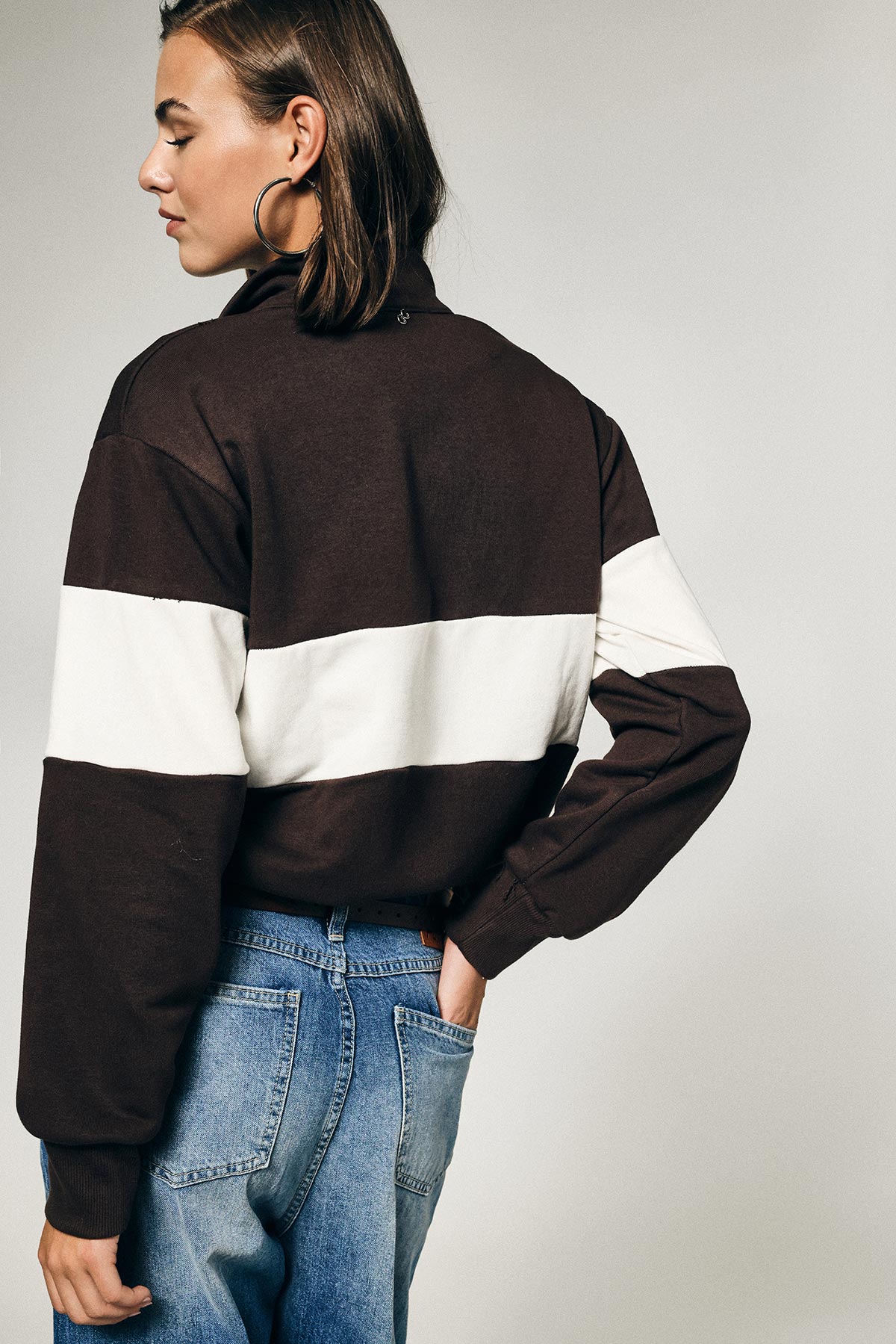 Woman seen from back and profile wearing brown jumper with white stripe, blue jeans and large hoop earring, hand in pocket
