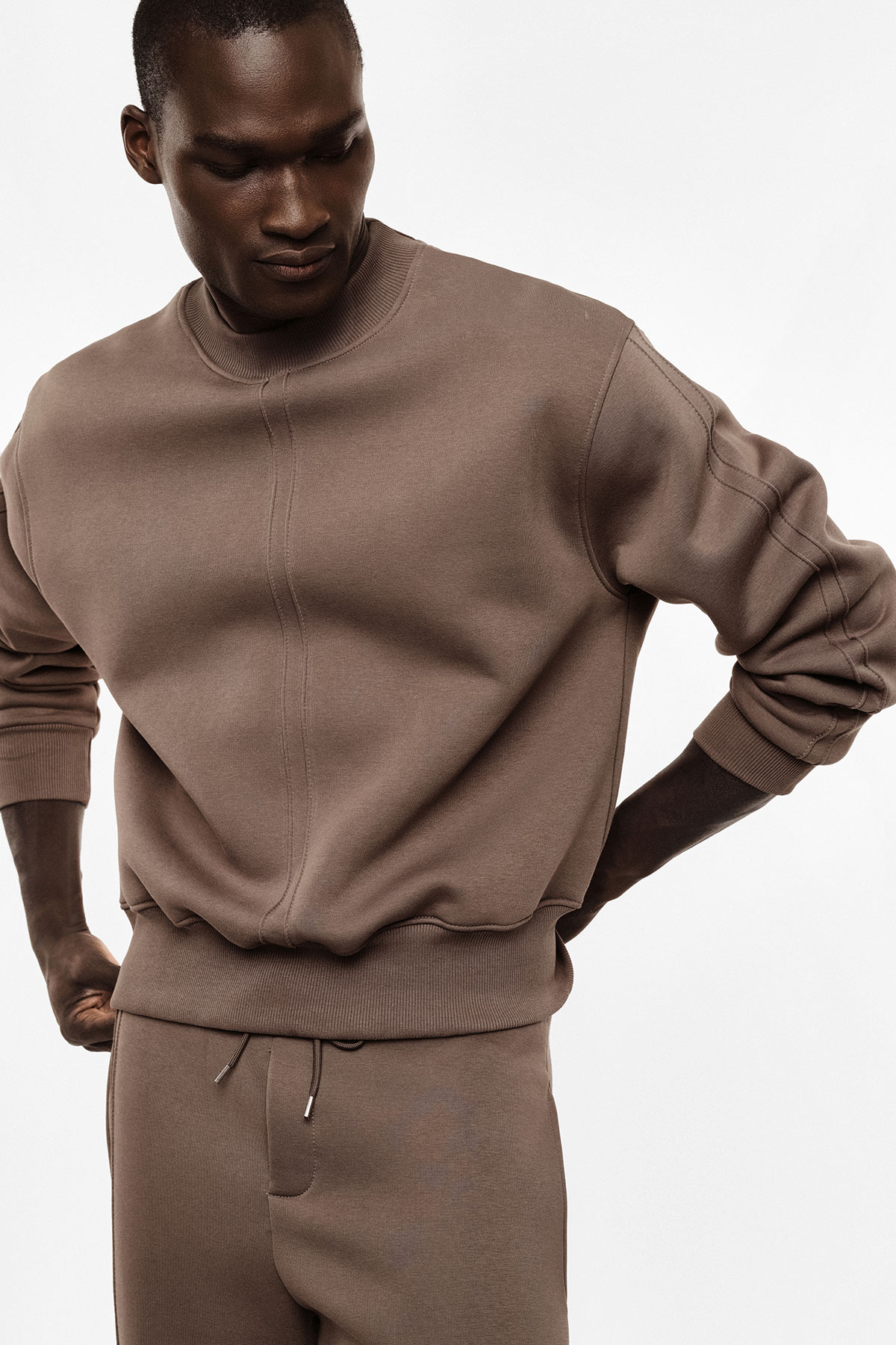 Male model wearing brown relaxed-fit sweatshirt and matching joggers with ribbed cuffs and hem, hands on hips
