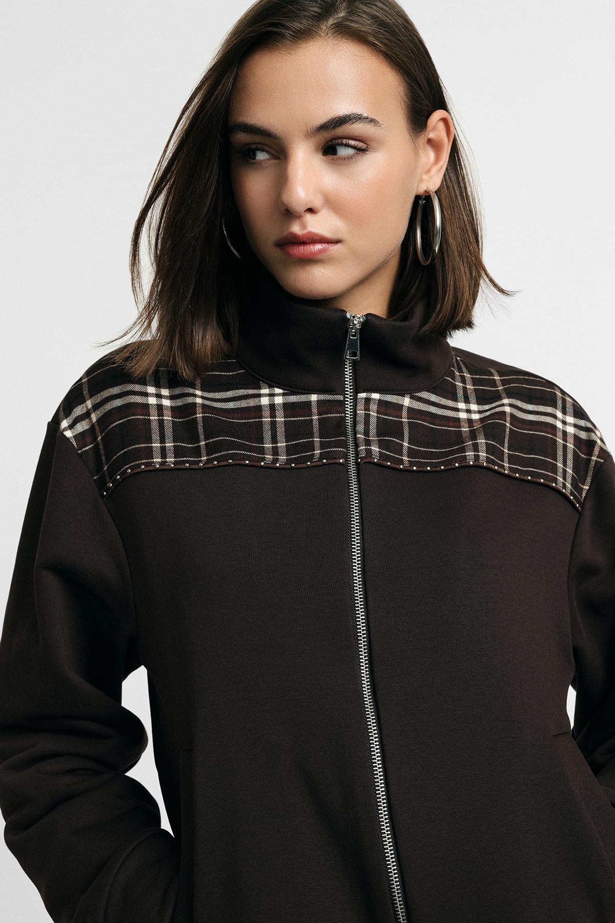Young woman wearing a dark brown zip-up jacket with plaid yoke and hoop earrings, looking to the side