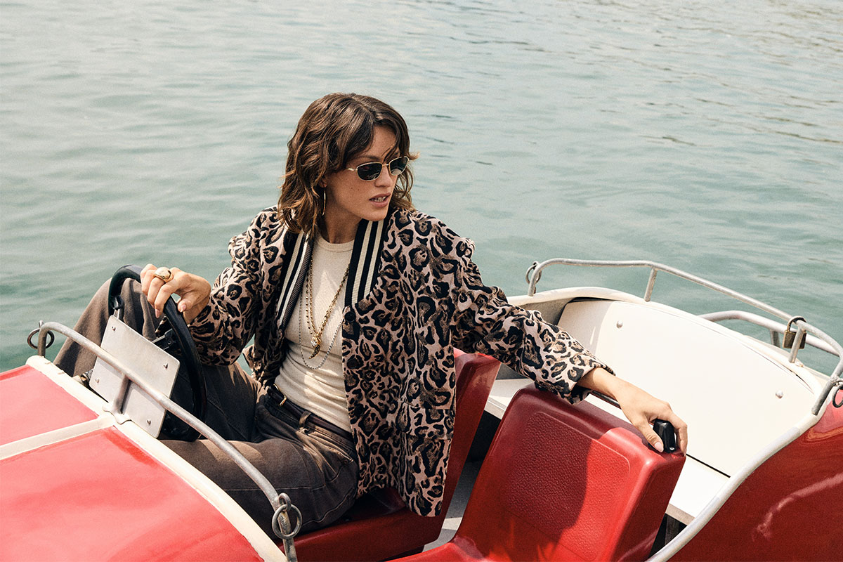 Woman in leopard-print jacket and sunglasses steering a red pedal boat on calm water, stylish jewellery visible