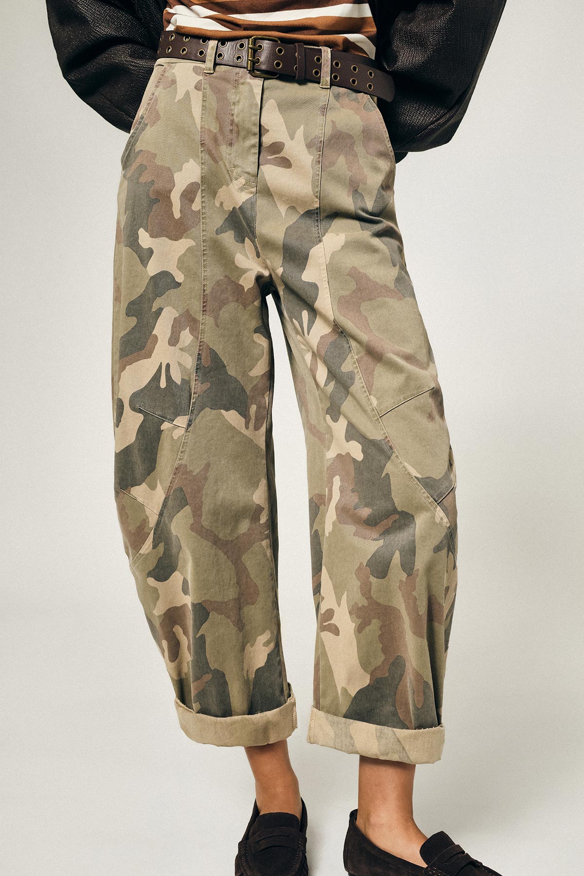 Cropped shot of person wearing camouflage cargo trousers with rolled cuffs, brown studded belt and black loafers