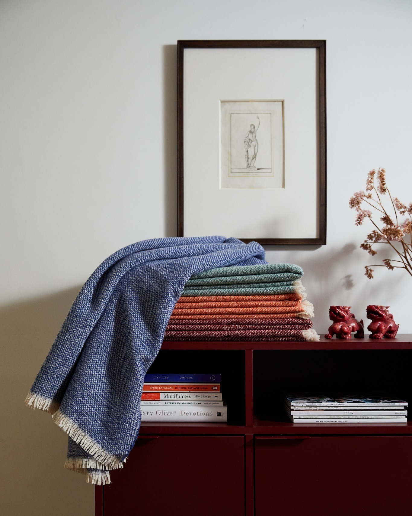 Blue throw draped over stack of colourful folded throws on a burgundy sideboard, with framed sketch, books and dried flowers