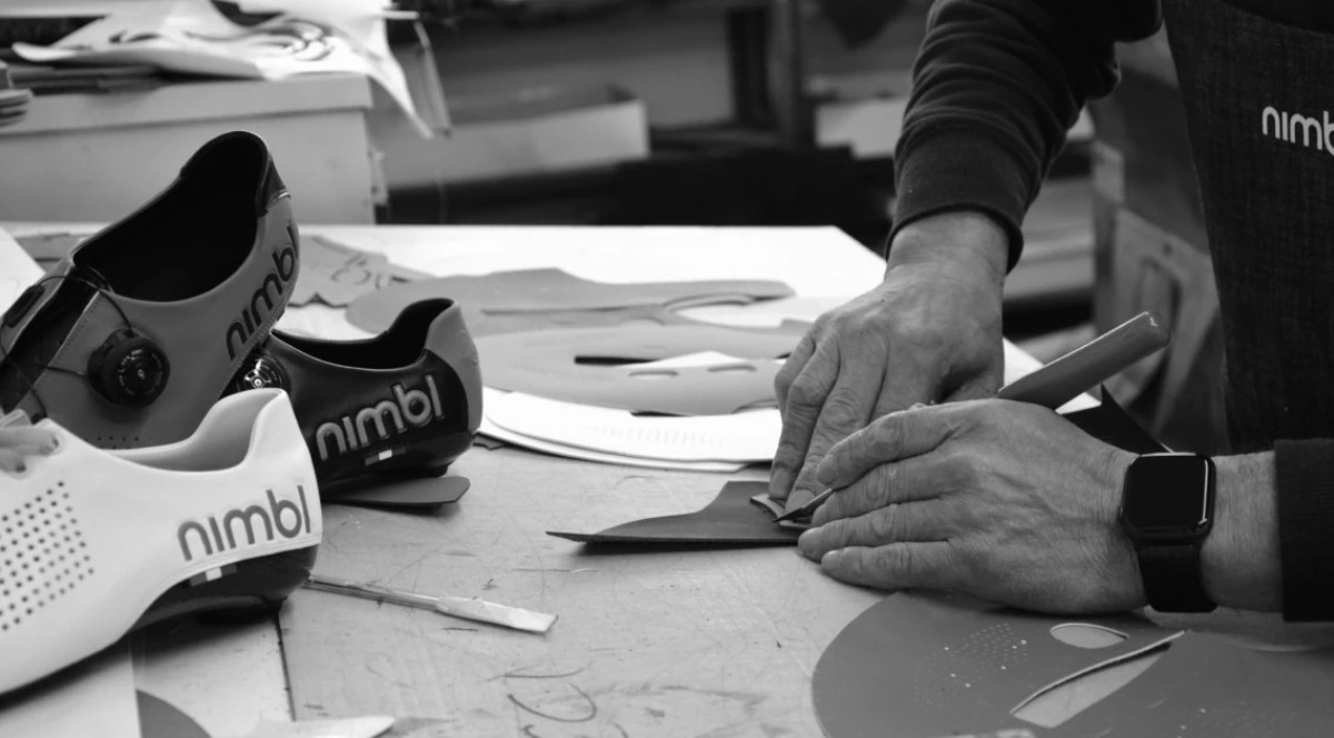 Hands cutting a leather pattern on a workbench beside white and black Nimbl cycling shoes during shoe crafting