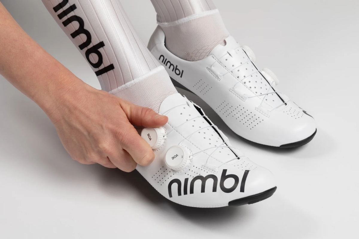 Close-up of white Nimbl road cycling shoes with dual BOA dials being adjusted by hand, wearer wearing white branded socks