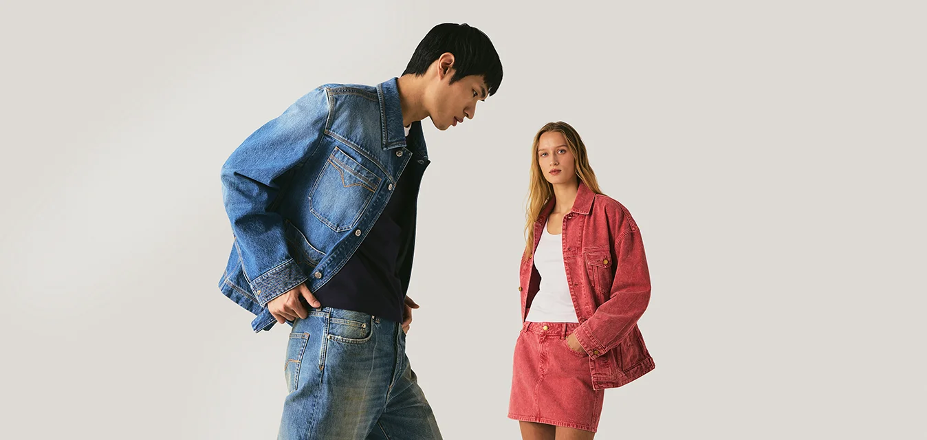 Giovane uomo con giacca di denim blu e jeans guarda verso il basso mentre donna con giacca di denim rossa e gonna guarda avanti su sfondo uniforme.
