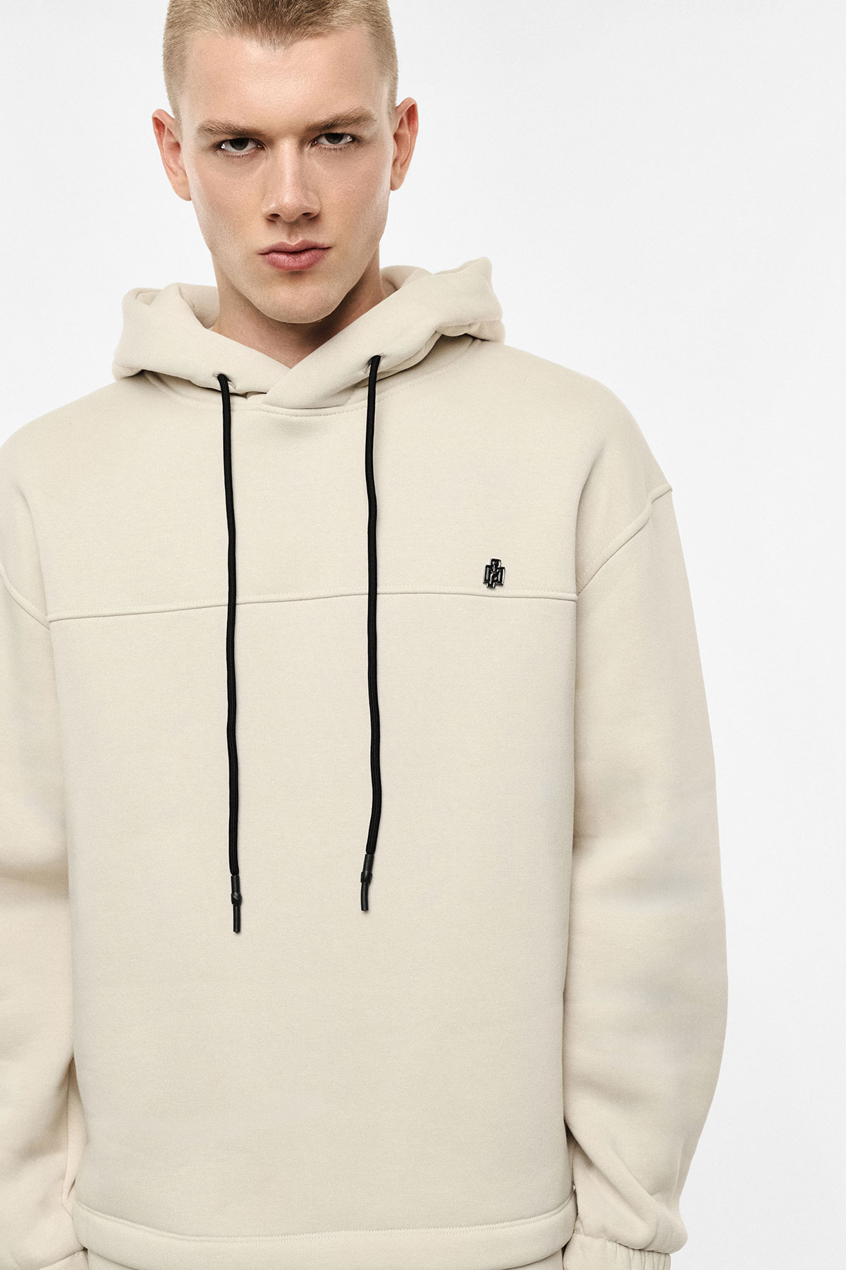 Young man wearing a beige hoodie with black drawstrings and small chest logo on a plain background