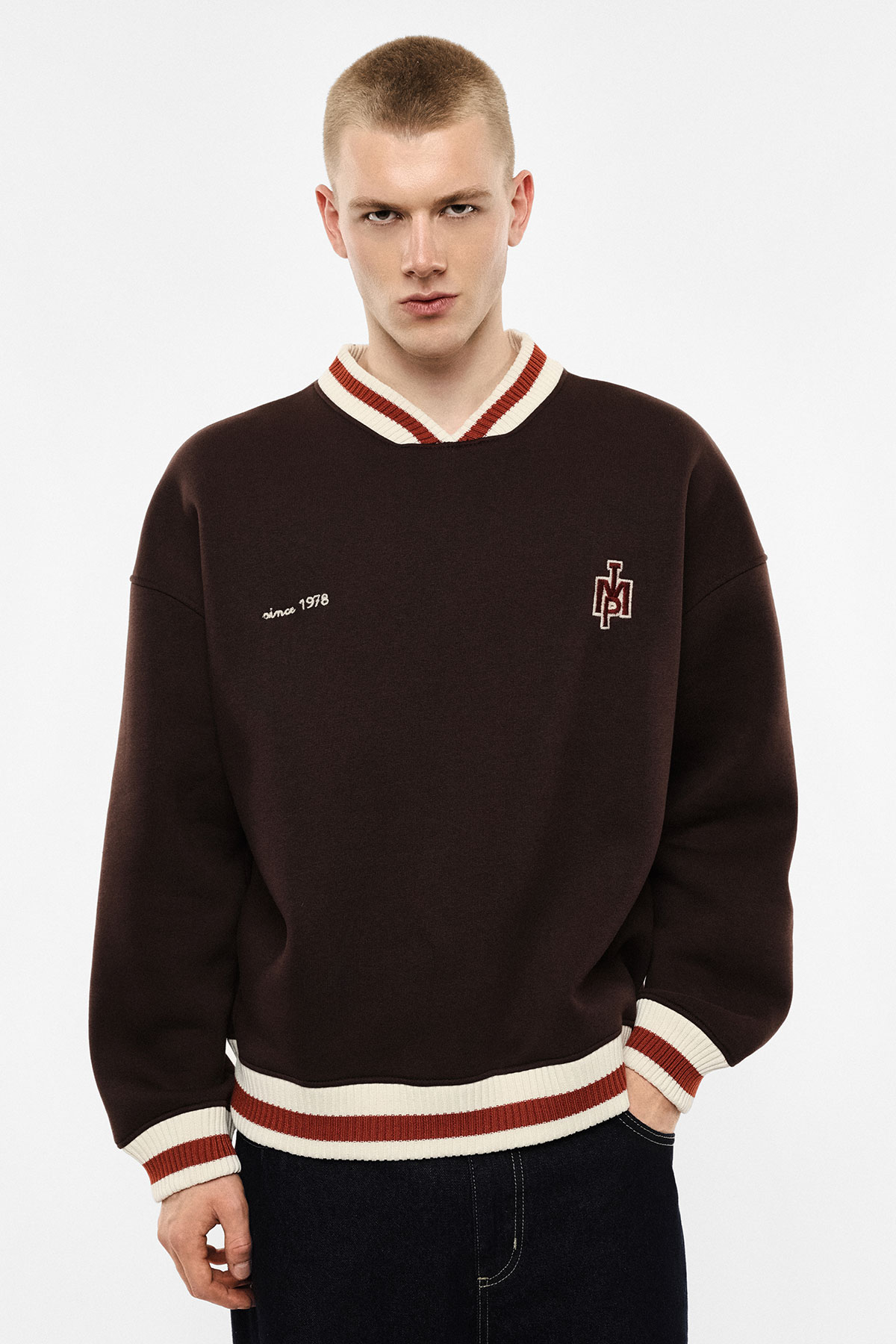 Young man in brown varsity-style sweater with cream and red striped collar and cuffs, embroidered logo and “since 1978” text on white background