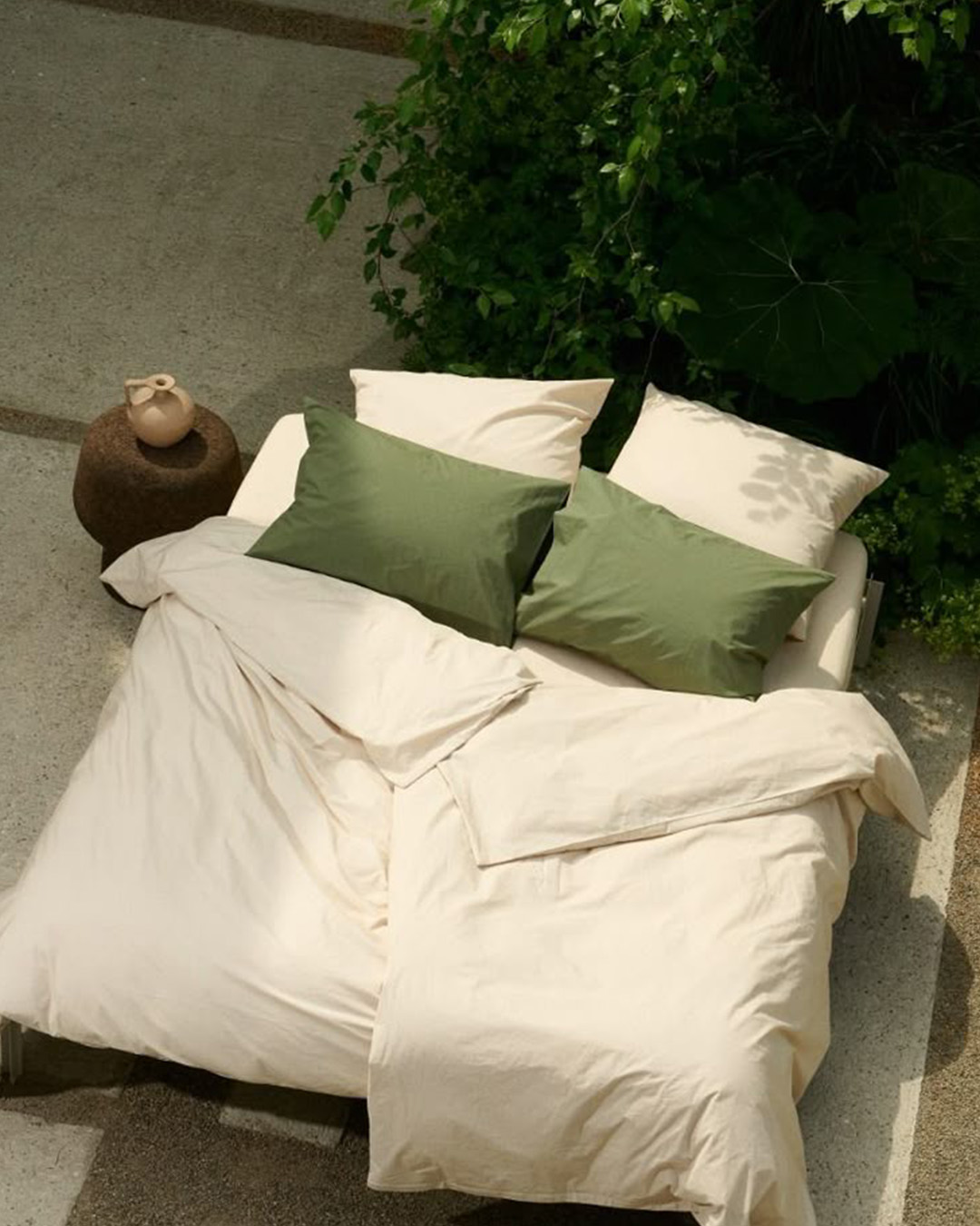 Cream duvet and sheets with two green pillowcases on an outdoor bed beside lush foliage and a small ceramic vase