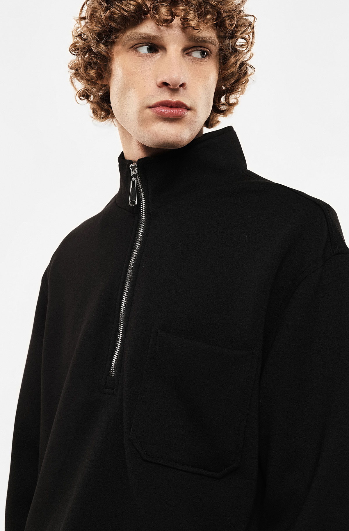 Young man with curly hair wearing black quarter-zip jumper with chest pocket, looking to the right on white background