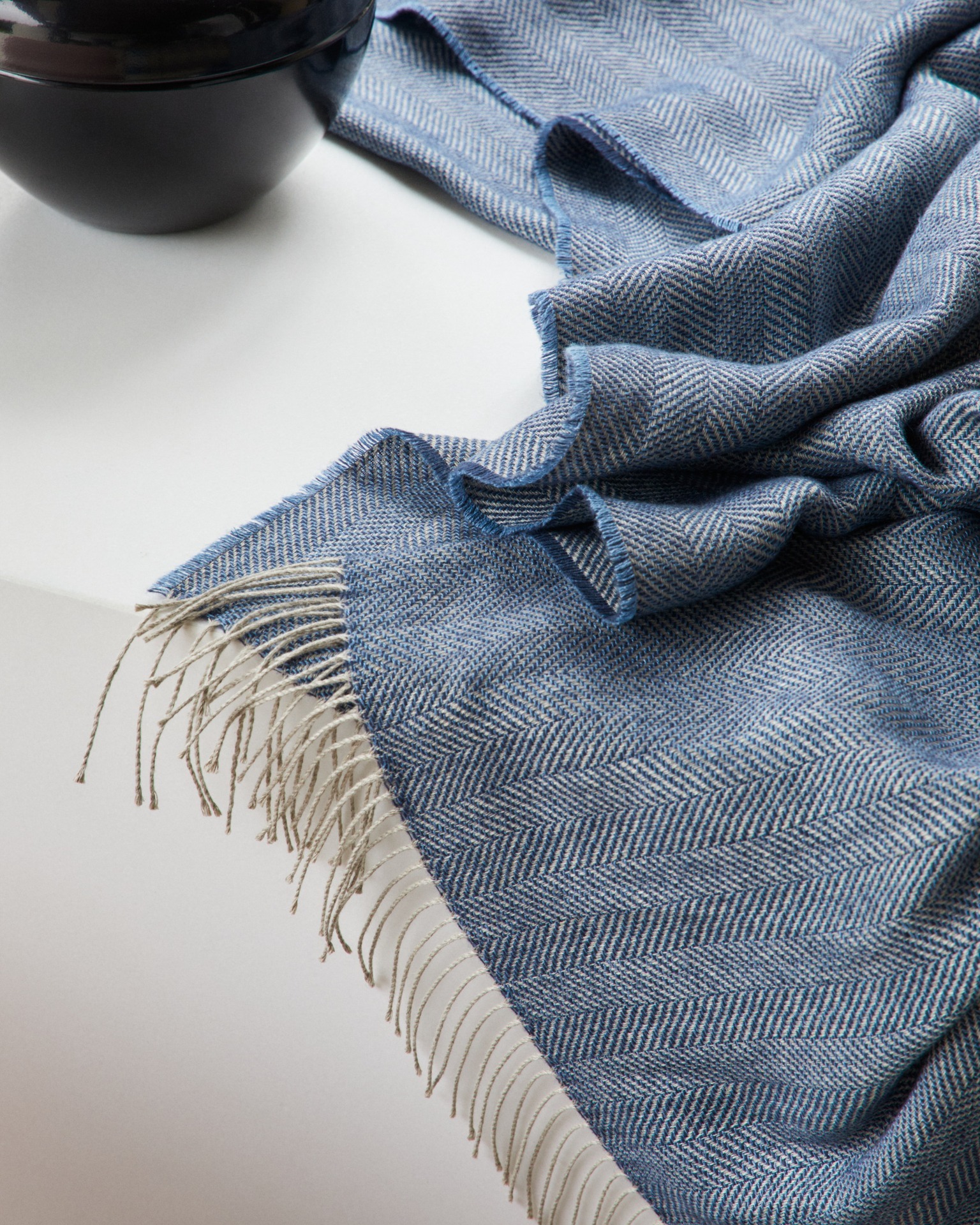Blue herringbone woven wool throw with beige fringe draped on white surface beside a black ceramic bowl