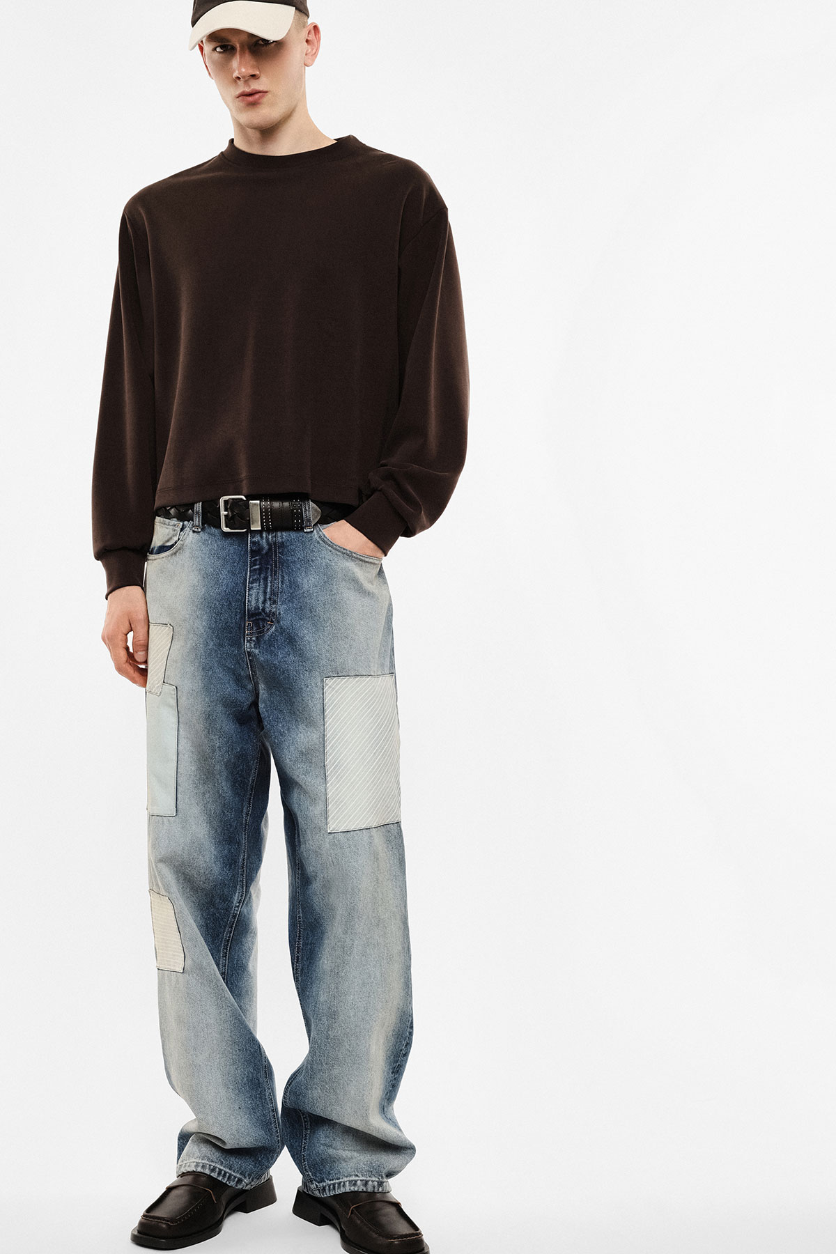 Male model wearing brown long-sleeve sweatshirt, patched loose blue jeans, black belt and shoes, beige cap on white background
