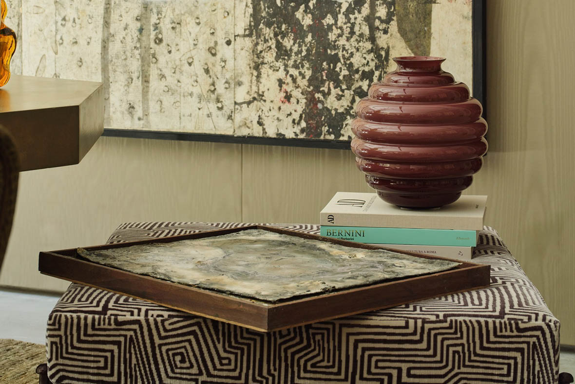 Modern living room vignette: patterned ottoman with wooden tray holding textured artwork, stacked books and maroon ribbed vase
