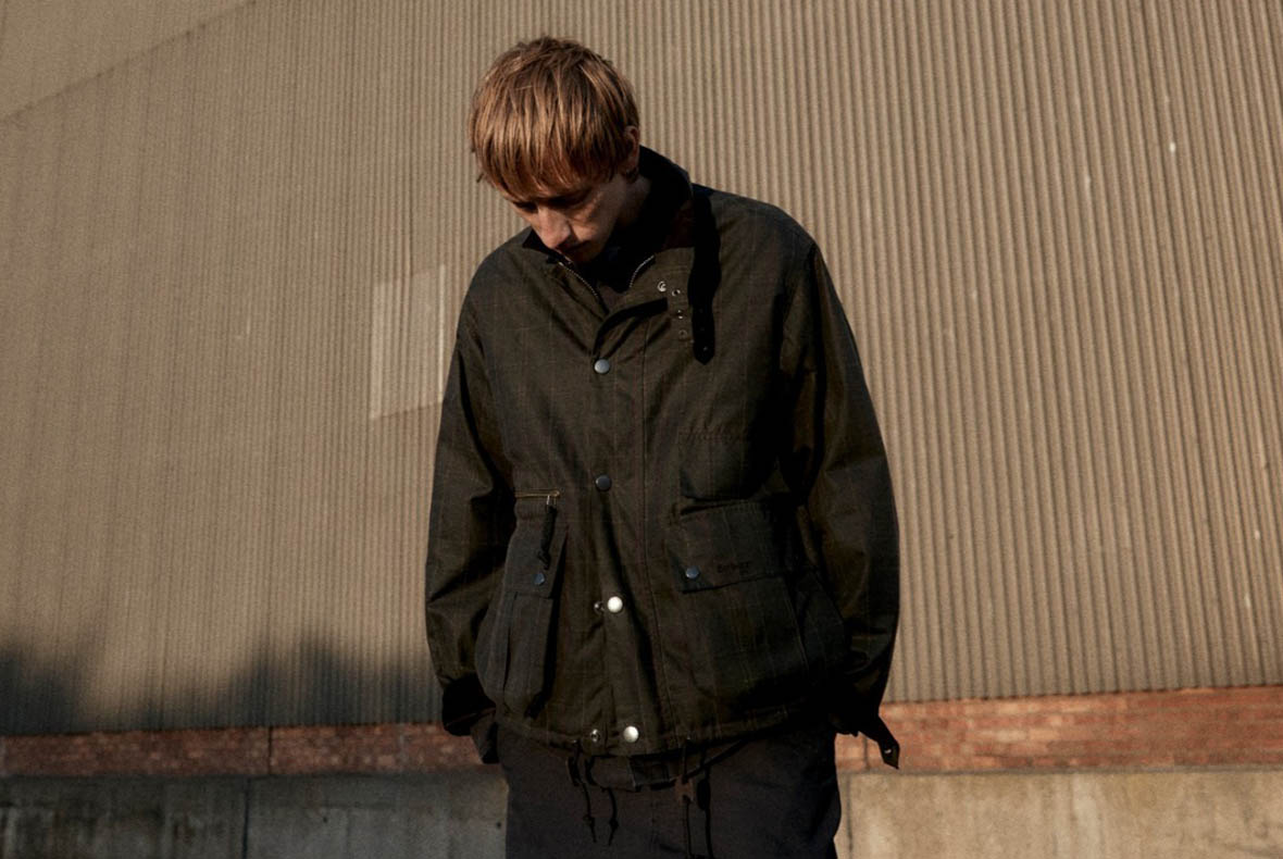 Young man wearing a dark jacket looking down in front of a corrugated metal wall, urban fashion portrait
