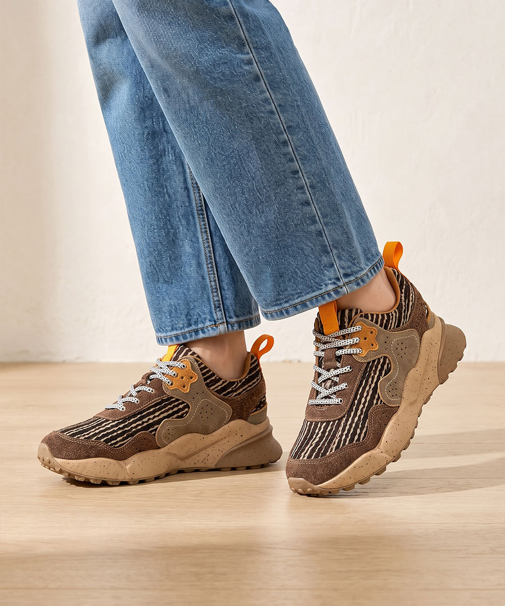 Pair of women's brown striped suede chunky sneakers with orange pull tabs and patterned laces, worn with cropped blue jeans on a wooden floor