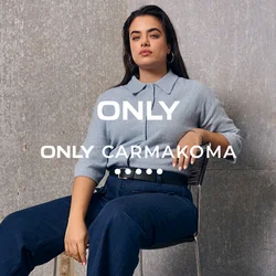 Donna con i capelli lunghi seduta su una sedia, indossa un maglione azzurro, pantaloni blu scuro e mocassini marroni, davanti a una parete industriale grigia.