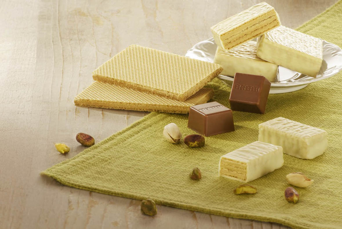 Assorted wafer biscuits and milk and white chocolate pralines with shelled pistachios on a green cloth and wooden table.