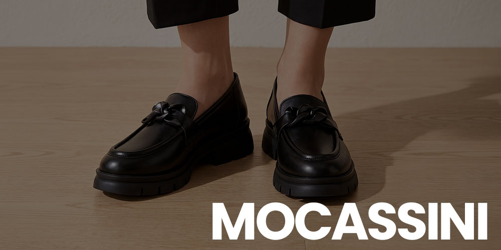 Black leather chunky-soled loafers with chain detail on wooden floor, model in cropped trousers; large 'MOCASSINI' brand text visible