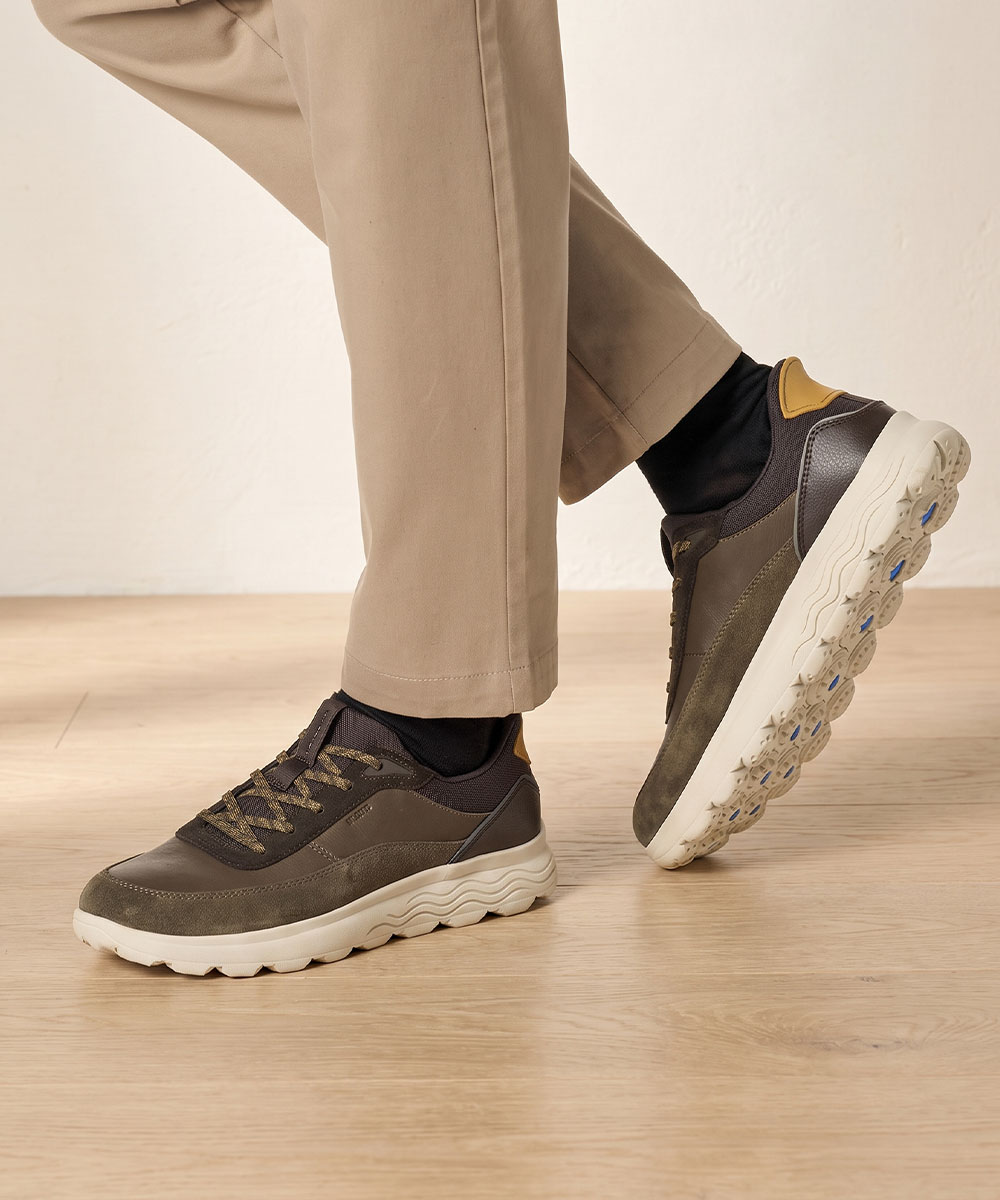 Close-up of men's brown trainers with chunky cream soles and patterned laces, worn with beige trousers on a wooden floor
