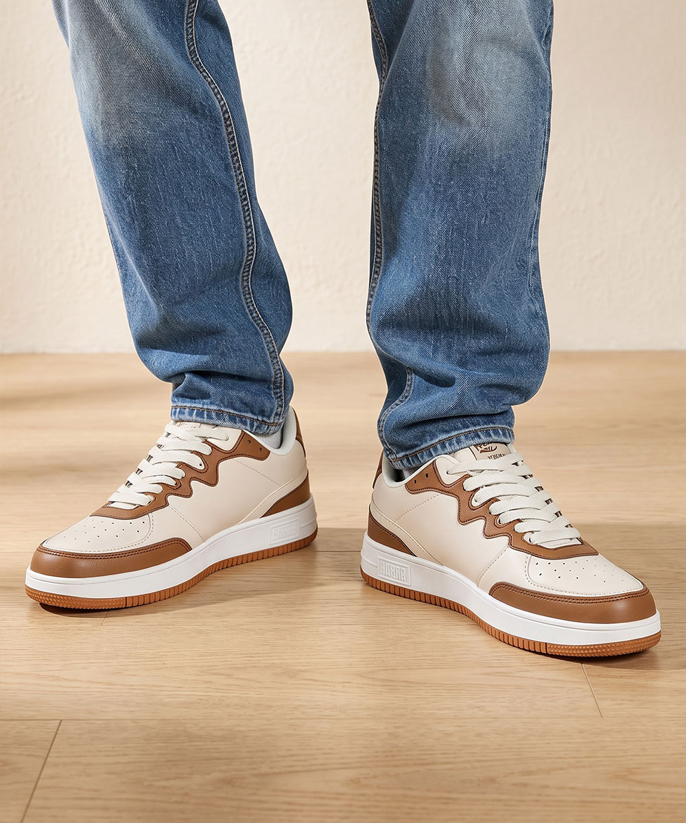 Pair of white and tan brown leather sneakers with gum soles worn with blue jeans, standing on light wooden floor