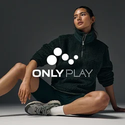Giovane donna con pullover in pile nero, pantaloncini neri, calzini bianchi e sneakers grigie seduta a terra davanti a uno sfondo scuro.