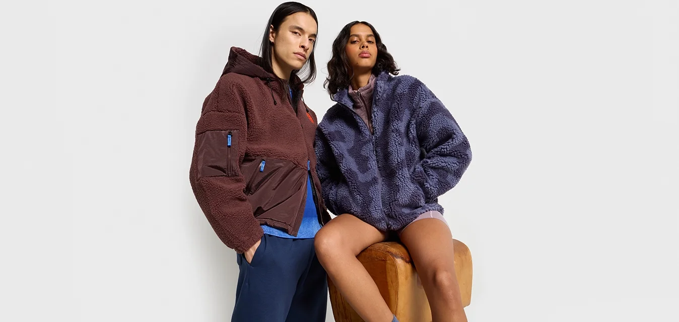 Giacca in pile marrone con zip e dettagli blu; giacca in pile viola con texture a motivo. Entrambi i modelli presentano cappucci e una vestibilità rilassata.
