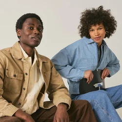 Due giovani adulti seduti su sedie contro uno sfondo semplice, uno con giacca marrone e beige, l’altro con un completo in denim blu e tacchi.