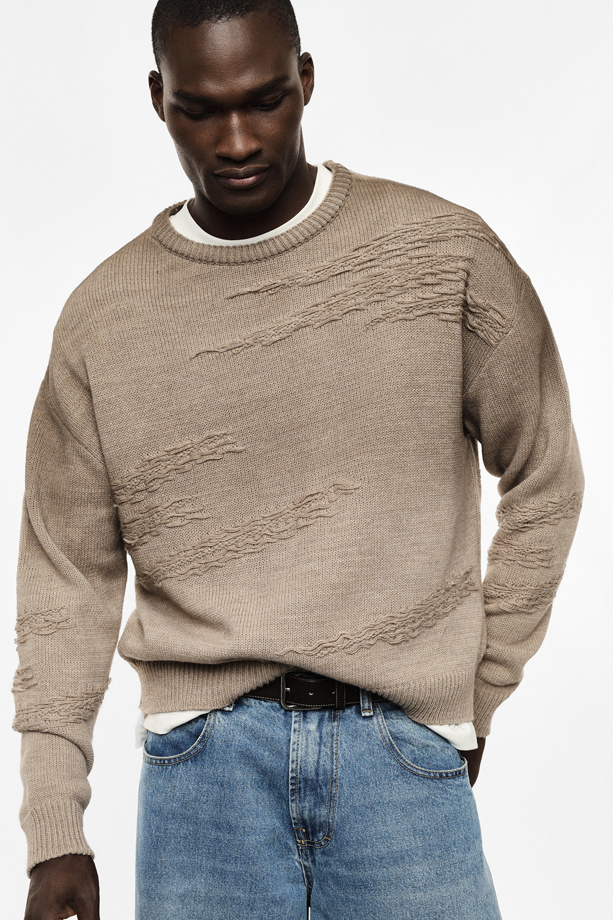 Male model wearing beige textured knit jumper over white T-shirt and blue jeans with black belt, looking down