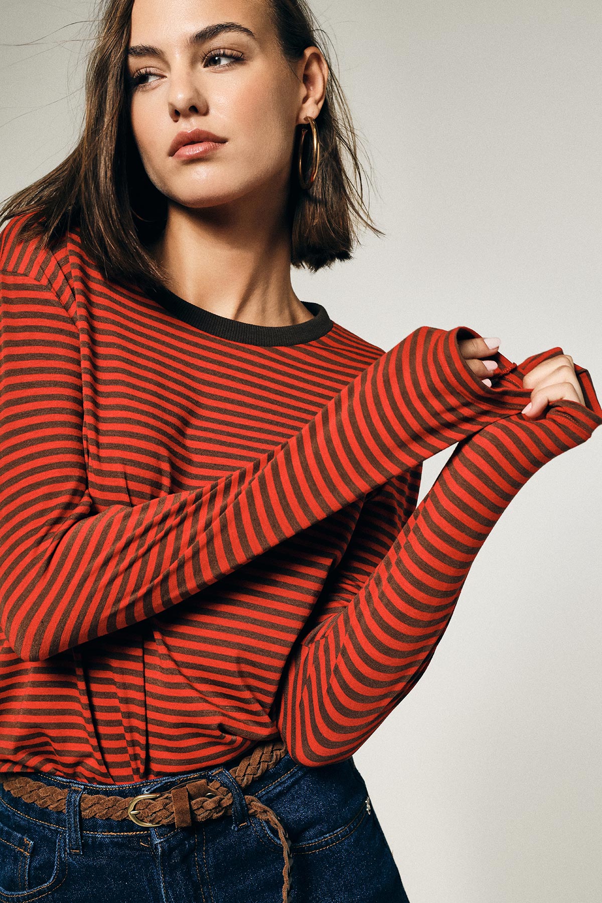 Young woman model wearing a red and black striped long-sleeve top, denim jeans and braided belt, hoop earrings, posing and looking left