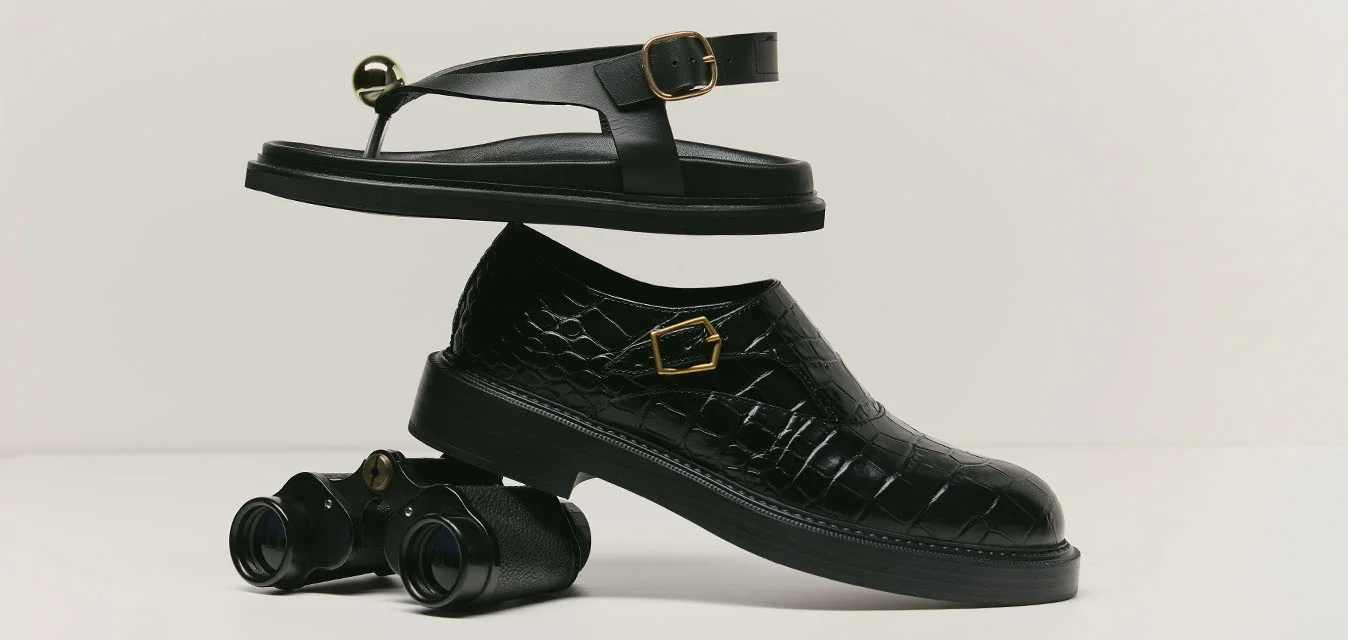 Sandalo nero bilanciato sopra una scarpa elegante nera con texture a coccodrillo appoggiata su un binocolo nero su sfondo bianco.