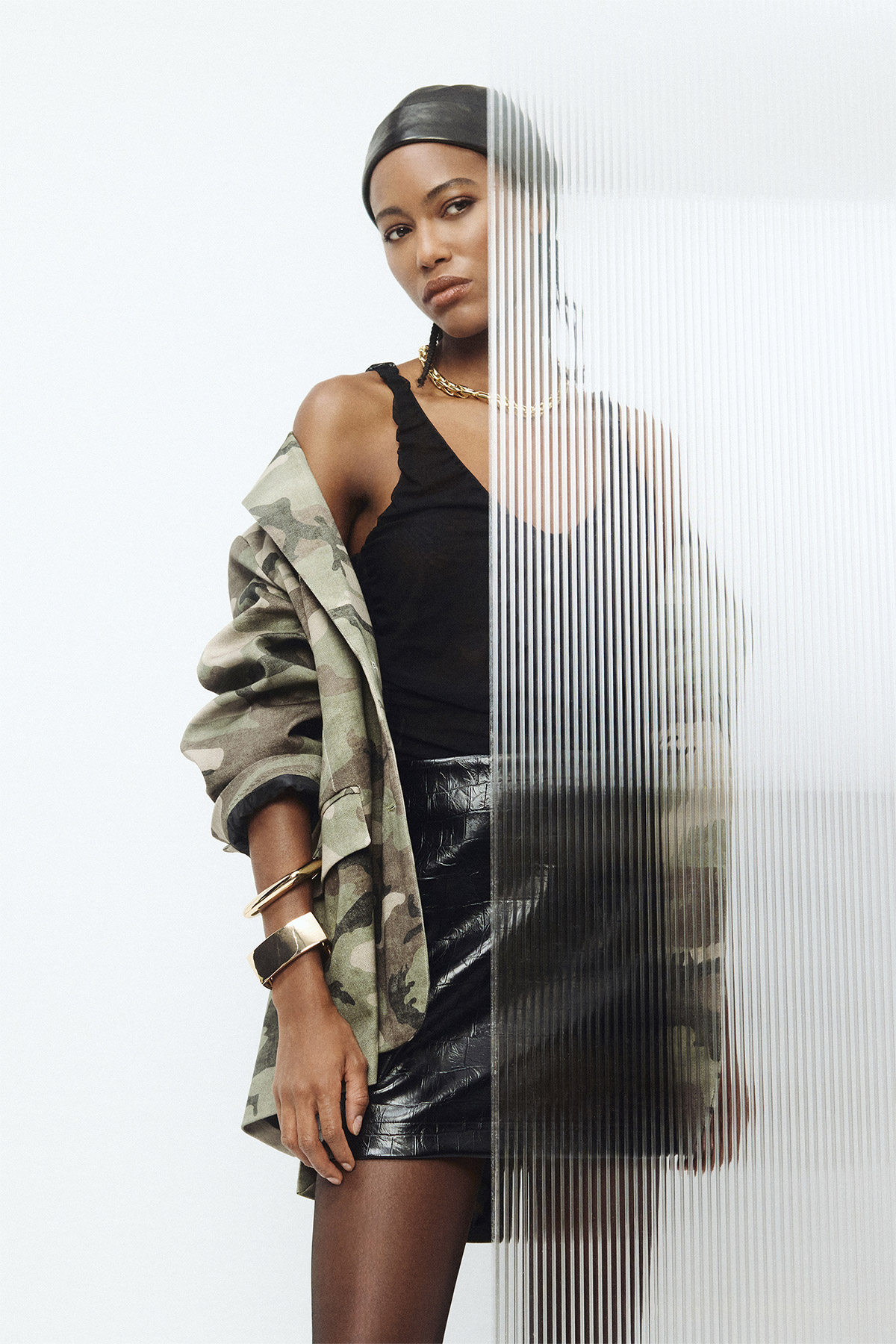 Female fashion model in camo jacket and black leather skirt behind ribbed glass panel, studio portrait, jewellery