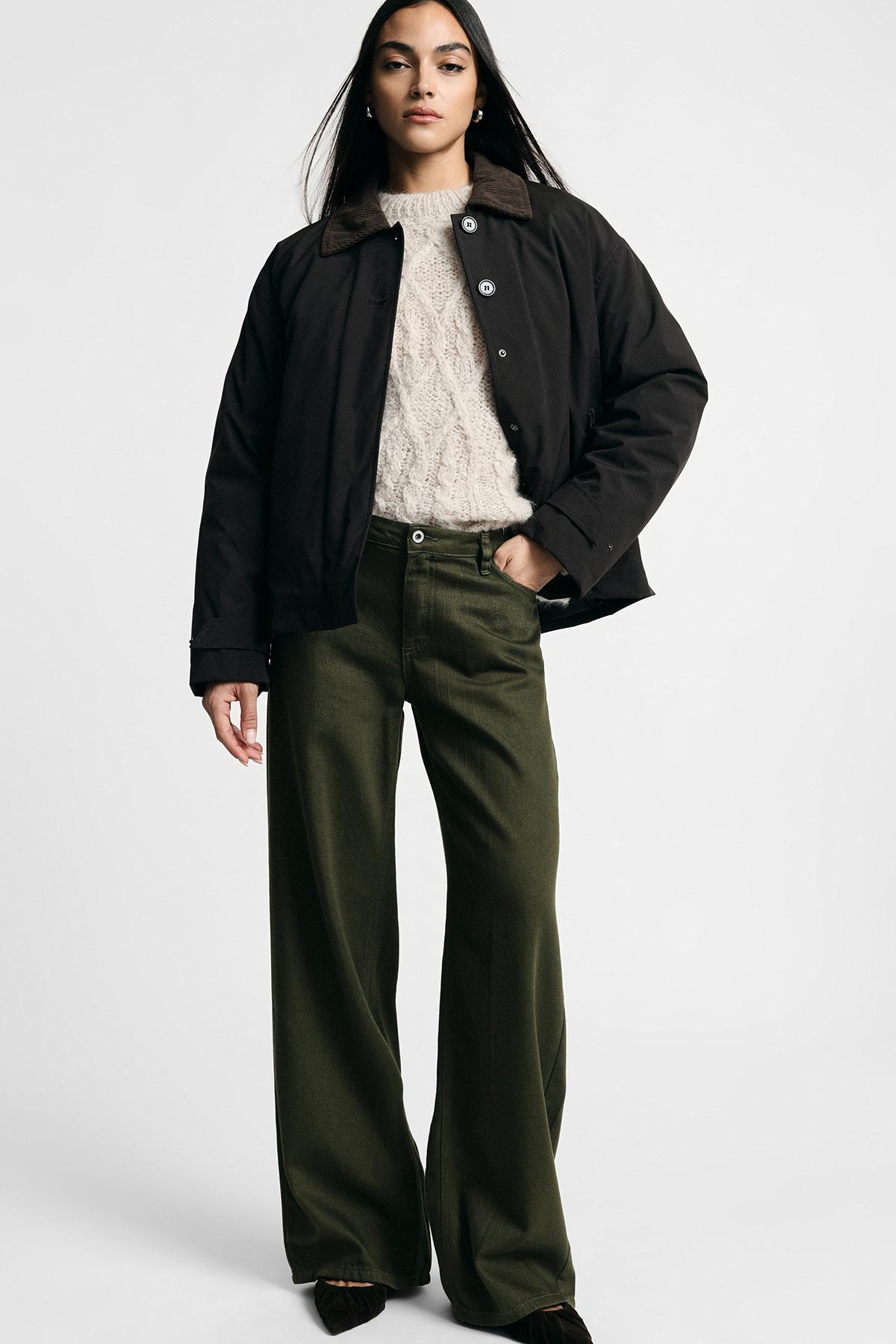 Woman standing against white background wearing a black jacket with cord collar, cream cable-knit sweater and olive wide-leg trousers
