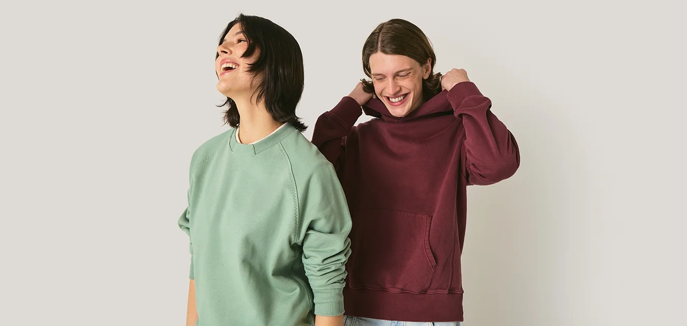 Felpa girocollo verde con polsini a coste indossata da una persona, affiancata da un hoodie bordeaux con tasca a canguro indossato da un'altra persona.