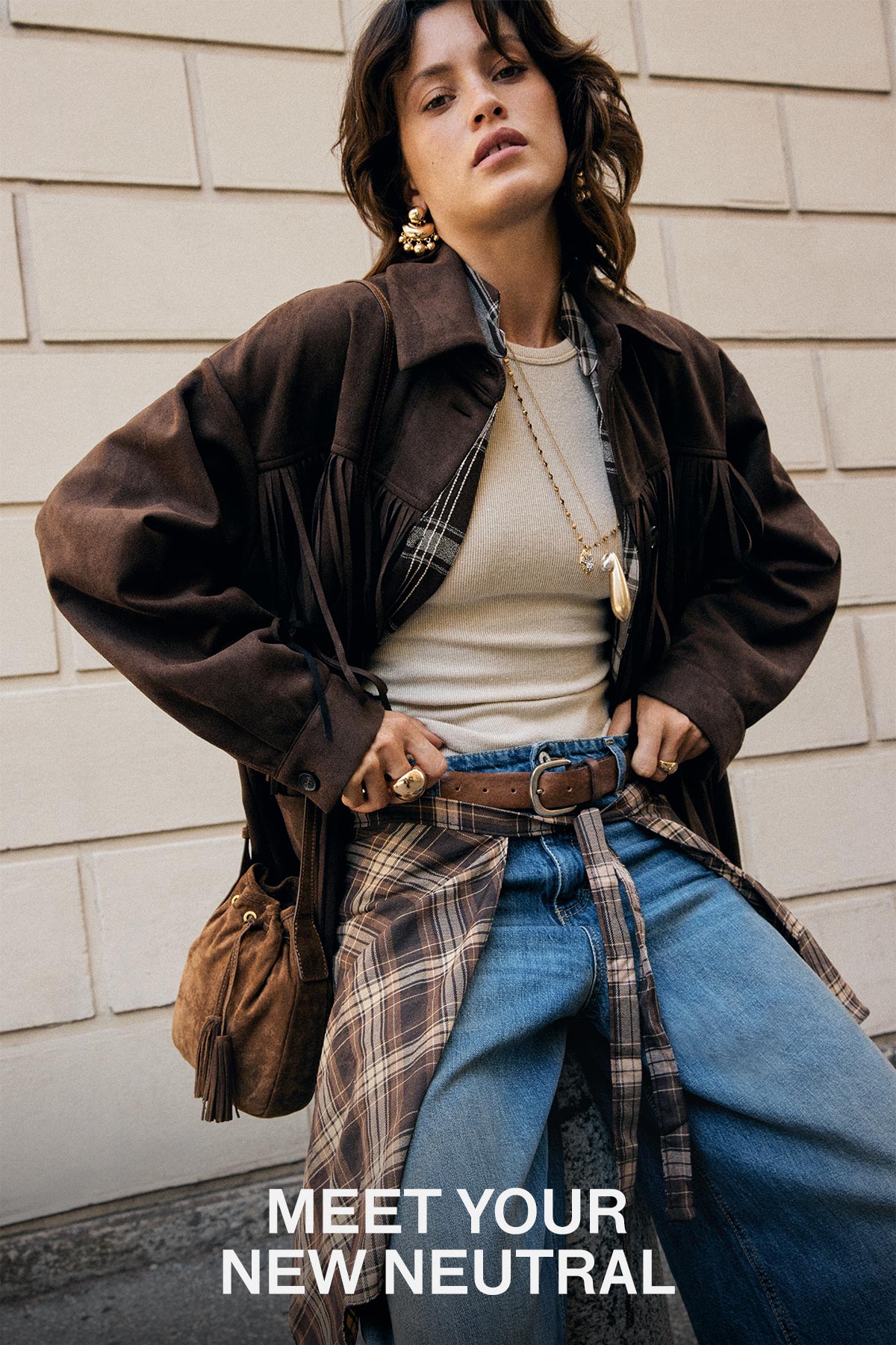 Woman in brown suede fringe jacket, cream ribbed top, layered necklaces and plaid shirt tied at waist over wide jeans, standing by tiled wall with text 'MEET YOUR NEW NEUTRAL'