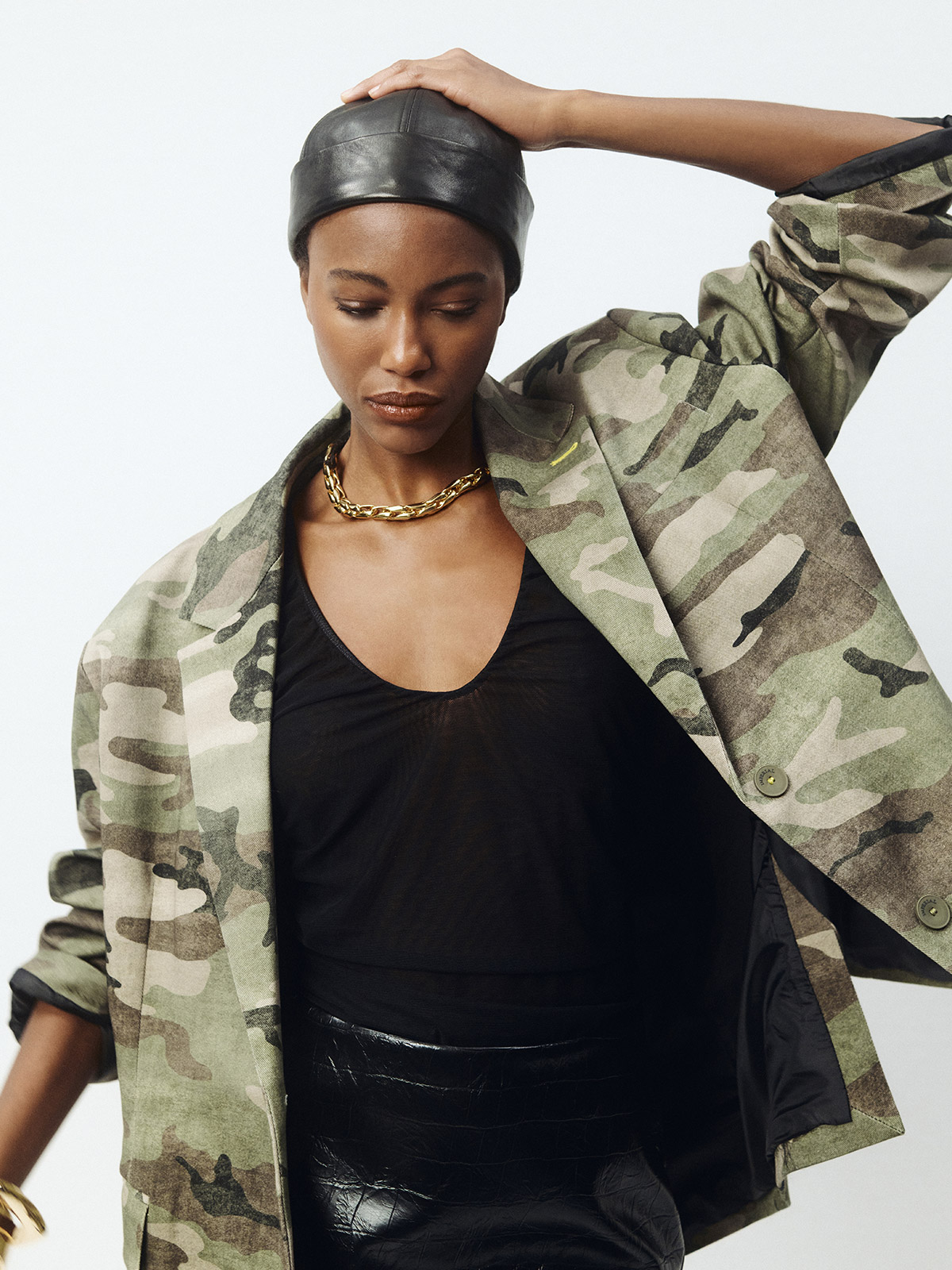 Model in camouflage jacket, black leather cap, gold chain necklace and sheer black top with glossy skirt in a fashion portrait