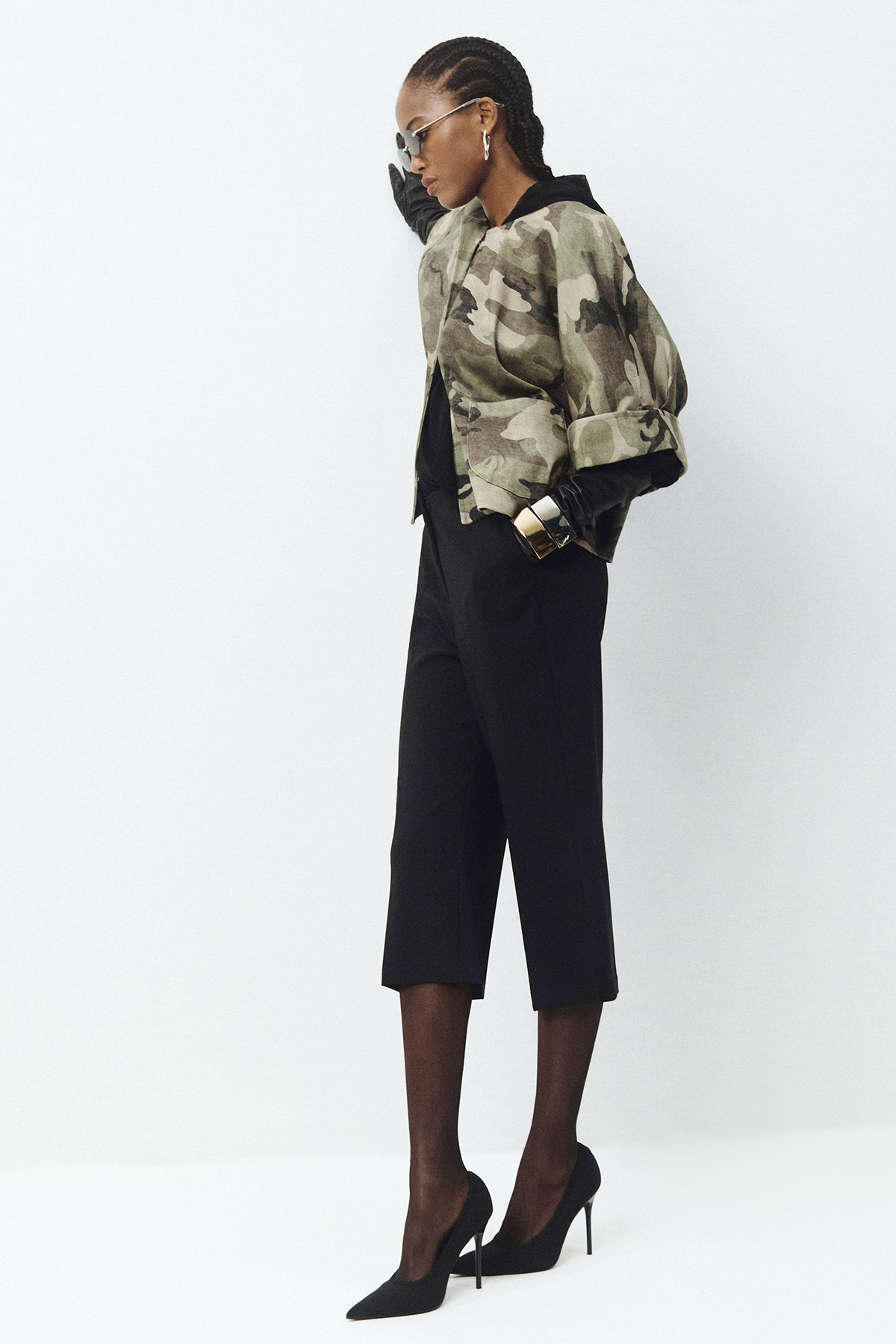 Model in profile wearing camo jacket, black cropped trousers and heels, sunglasses and gold cuff on white background