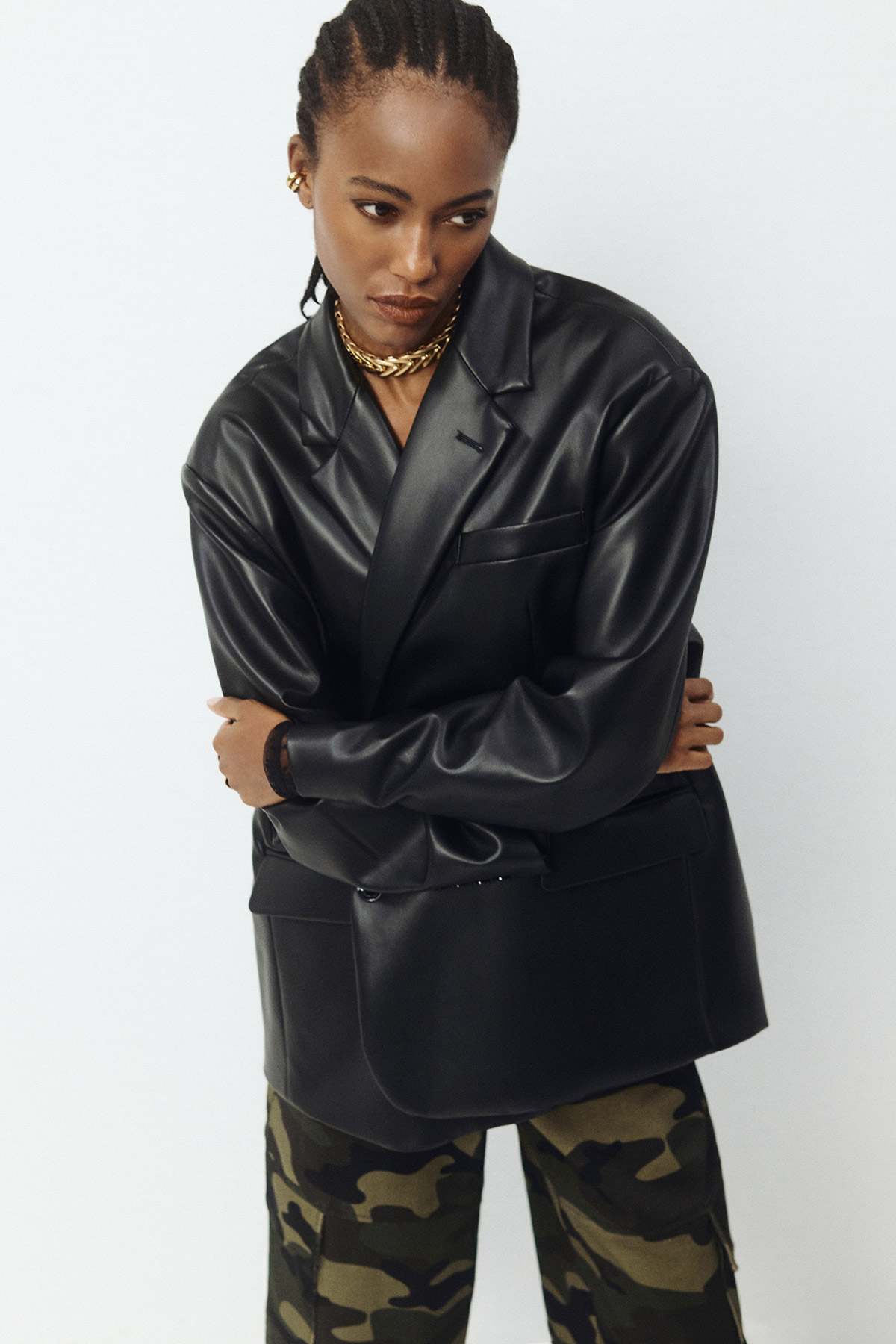 Model wearing black faux-leather blazer, camo cargo trousers and chunky gold jewellery, arms crossed, looking down.