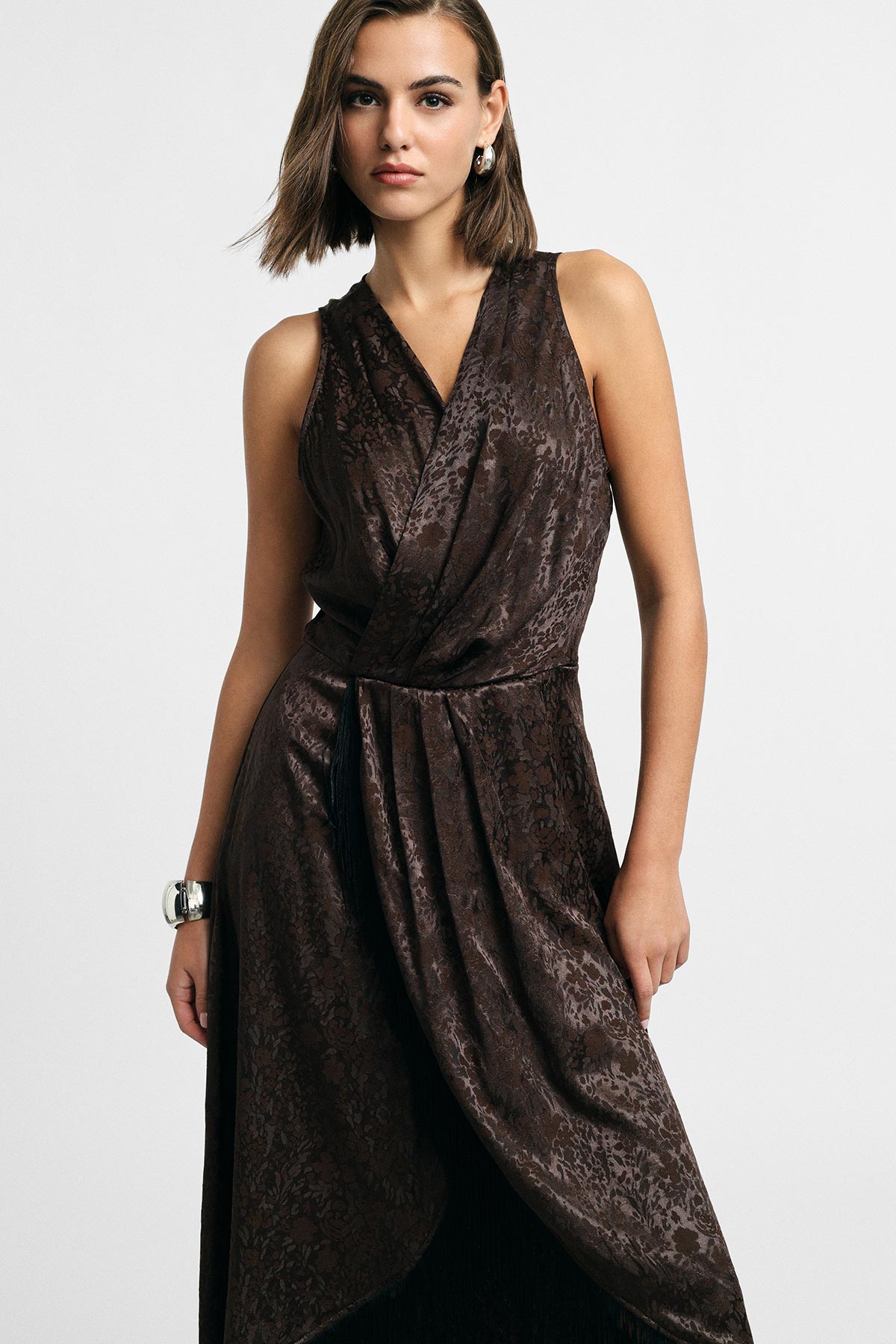 Model wearing a sleeveless brown patterned wrap dress with draped skirt, short bob hair, silver cuff bracelet and hoop earring on plain background